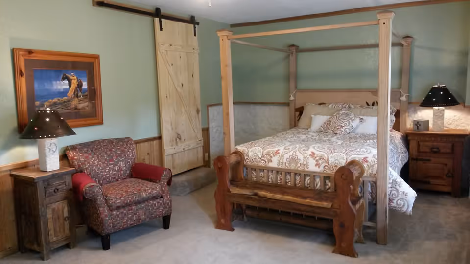A cozy bedroom featuring a wooden four-poster bed with patterned bedding, a rustic wooden bench at the foot of the bed, two wooden nightstands with lamps, a patterned armchair with red armrest covers, a framed picture on the wall, and a sliding barn door.