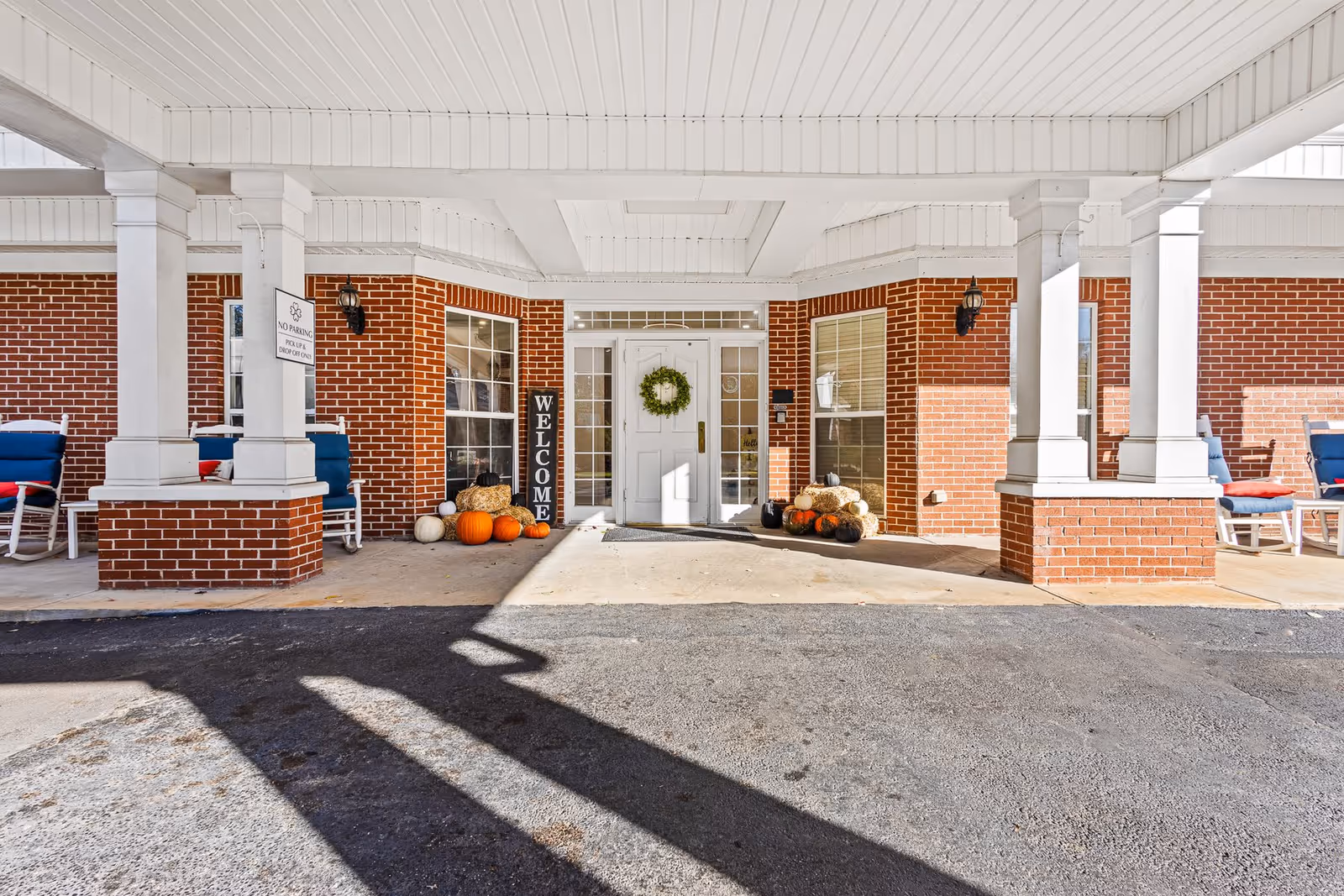 Entrance of a brick building with white columns and a covered porch. The door is white with glass panels and decorated with a green wreath. There are pumpkins and hay bales arranged on either side of the door, along with a vertical welcome sign. Blue rocking chairs with red cushions are placed on the porch.
