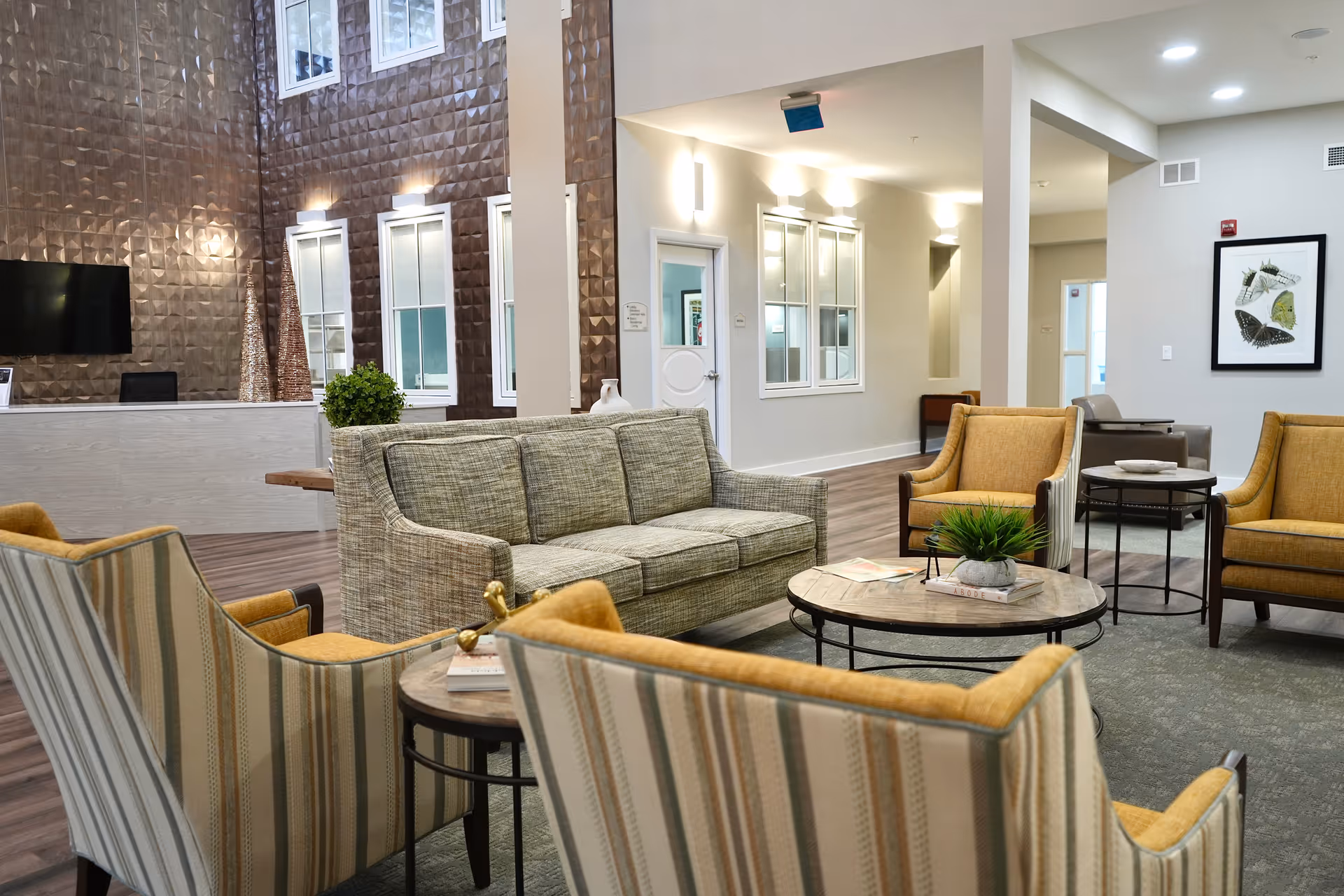 A modern and cozy living room area with a green sofa, four yellow and striped armchairs arranged around two round coffee tables. The room has a textured brown accent wall with windows and a mounted TV, light-colored walls, and wood flooring. There are decorative plants and framed artwork on the walls.