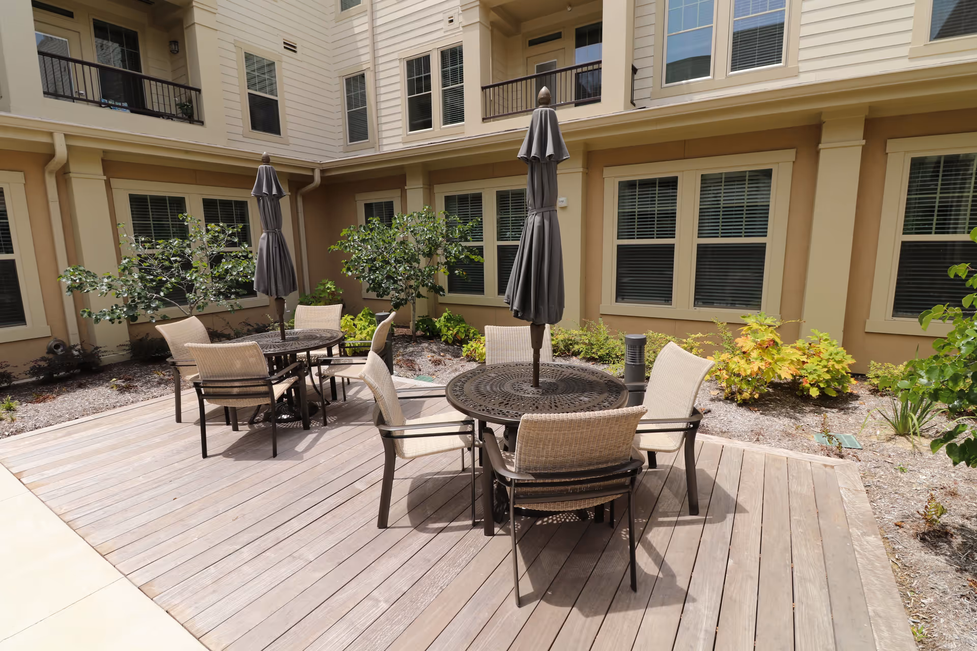 Outdoor patio area at an assisted living facility with two round metal tables, each surrounded by four beige chairs. Each table has a closed dark umbrella in the center. The patio is made of wooden decking and is adjacent to a beige building with multiple windows and some landscaping with small bushes and plants.