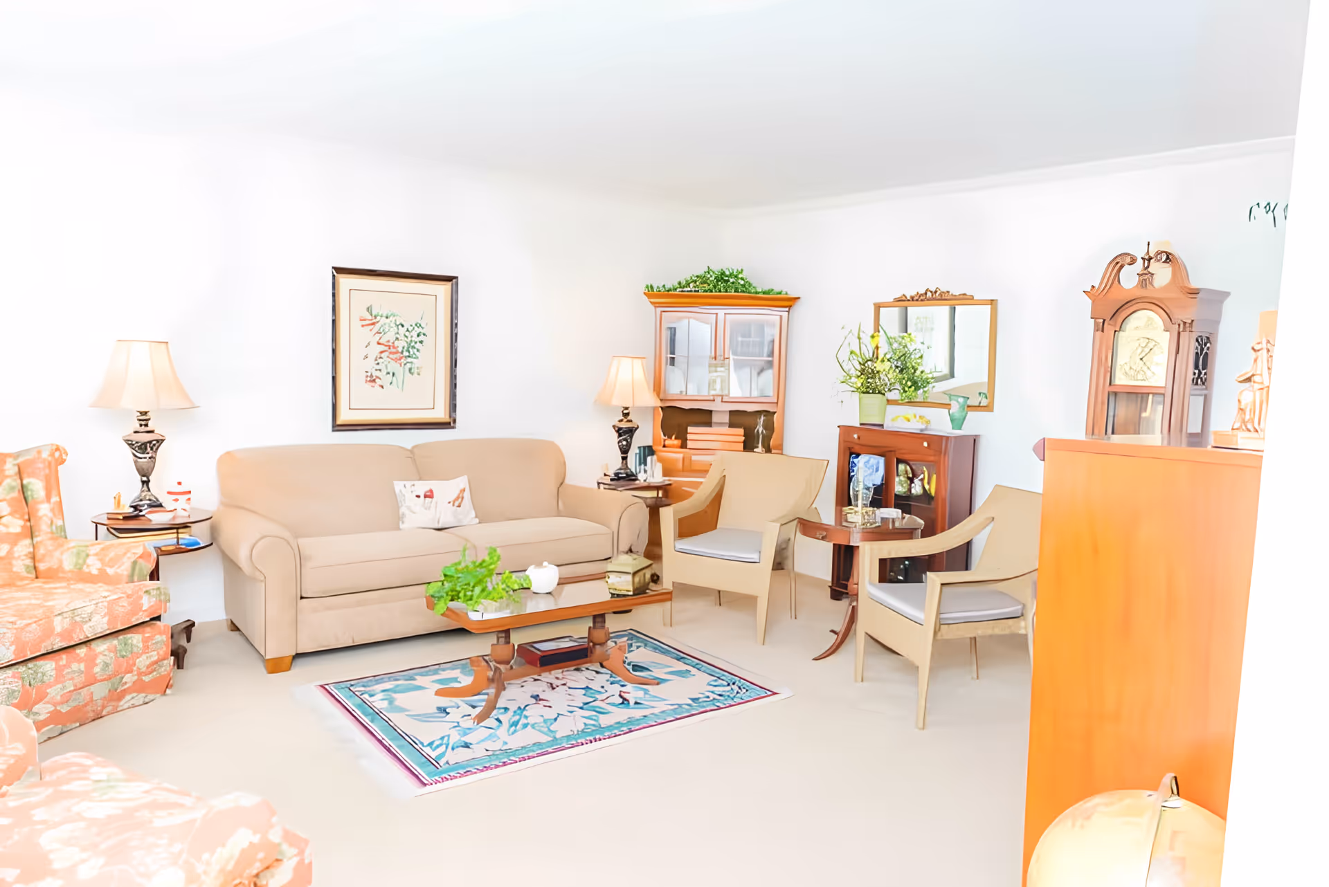 A bright and cozy living room with a beige sofa, two beige armchairs, and two floral-patterned armchairs. There is a wooden coffee table with a plant and decorative items on a floral rug. The room also features two side tables with lamps, a wooden cabinet with glass doors, a mirror above a small wooden cabinet with plants, and a tall grandfather clock. The walls are white and decorated with framed artwork.