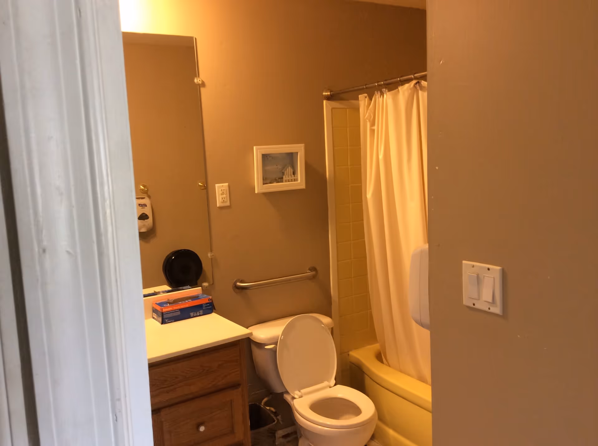 Small beige bathroom showing a vanity with mirror, toilet, and a bathtub with a shower curtain and grab bar.