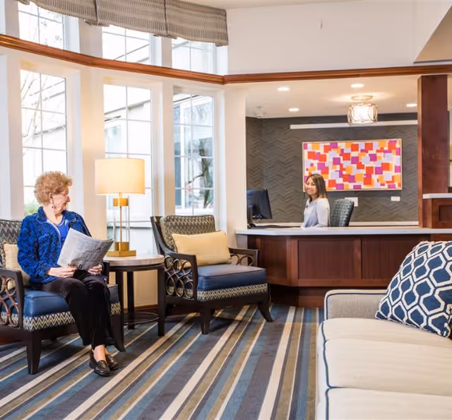 A senior woman sitting on a chair reading a newspaper in a well-lit reception area with large windows. A receptionist is seated behind a wooden desk with a colorful abstract painting on the wall behind her. The room has patterned chairs, a striped carpet, and a beige sofa with a geometric patterned pillow.