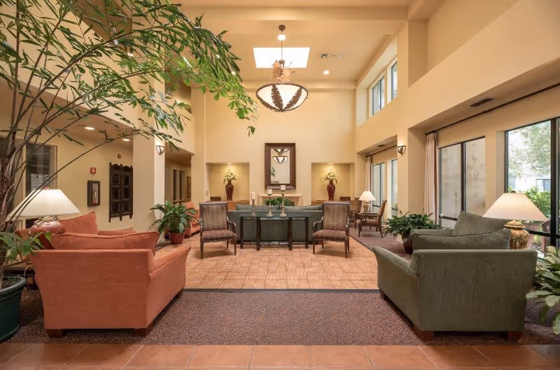 A bright and spacious senior living facility common area with high ceilings and large windows. The room features comfortable seating including a red sofa on the left and a green sofa on the right, several armchairs, potted plants, table lamps, and a central table with decorative items. A large hanging light fixture and a skylight provide ample lighting, and a mirror and vases are displayed on the far wall.