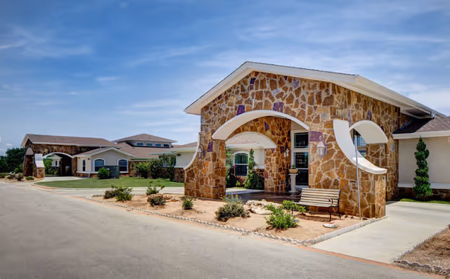 Stone-faced assisted living facility exterior with curved arches, benches, and landscaped entrance.