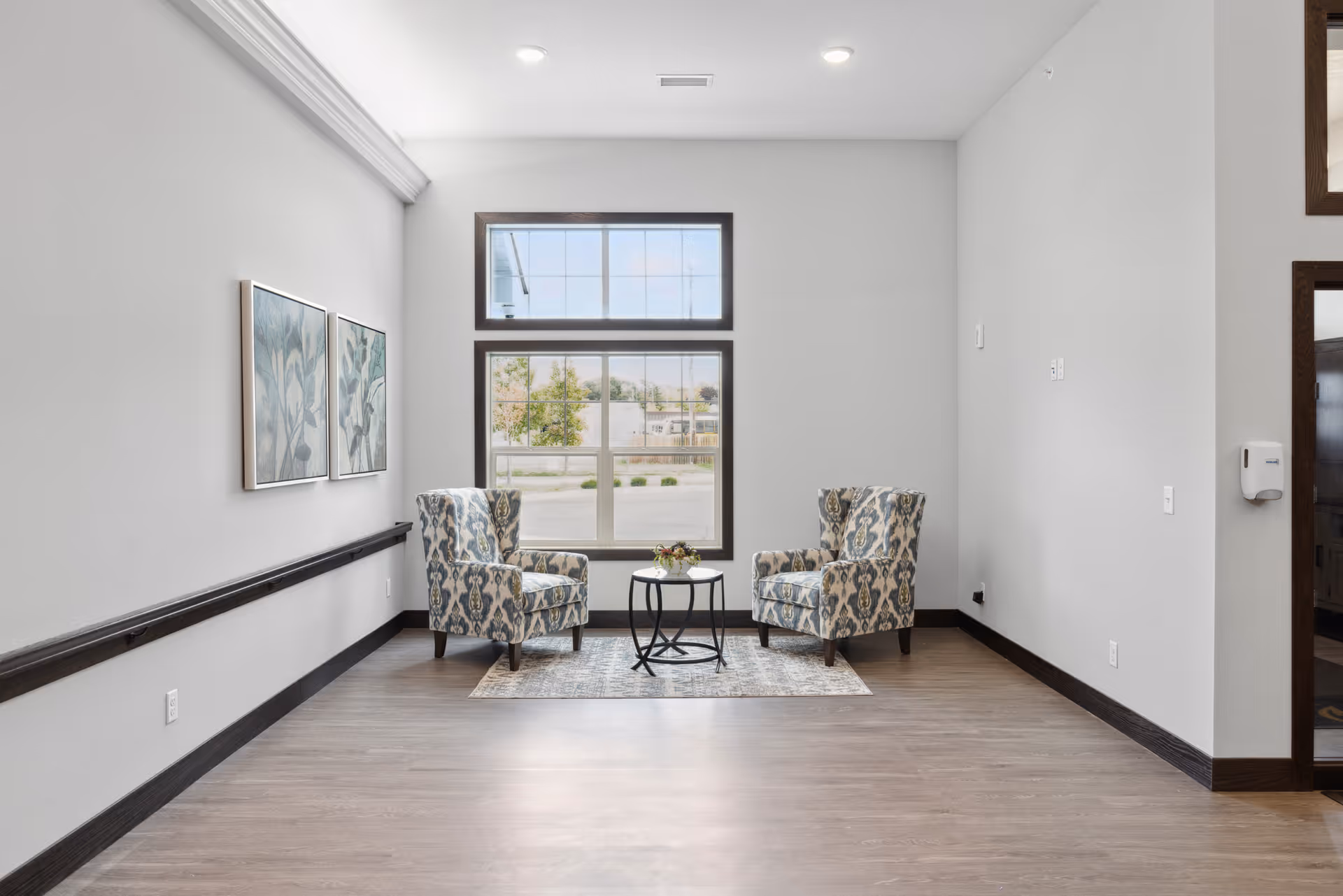 A small seating area in a senior living facility with two patterned armchairs facing each other across a small round table on a rug. The room has light gray walls, a large window with two panes letting in natural light, two framed botanical prints on the left wall, and wood-look flooring.