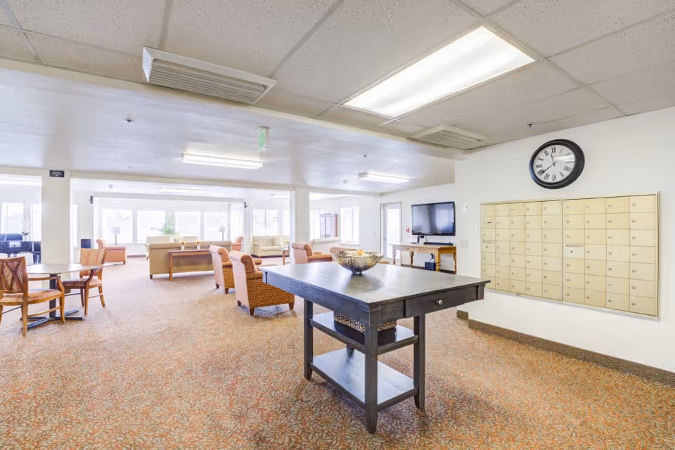 A spacious common area in a senior living facility with multiple seating arrangements including armchairs and sofas. There is a large black table in the center with a decorative bowl on top. A wall-mounted clock and a set of mailboxes are visible on the right wall. The room is well-lit with ceiling lights and natural light from large windows in the background. A flat-screen TV is mounted on the wall near a small wooden table.