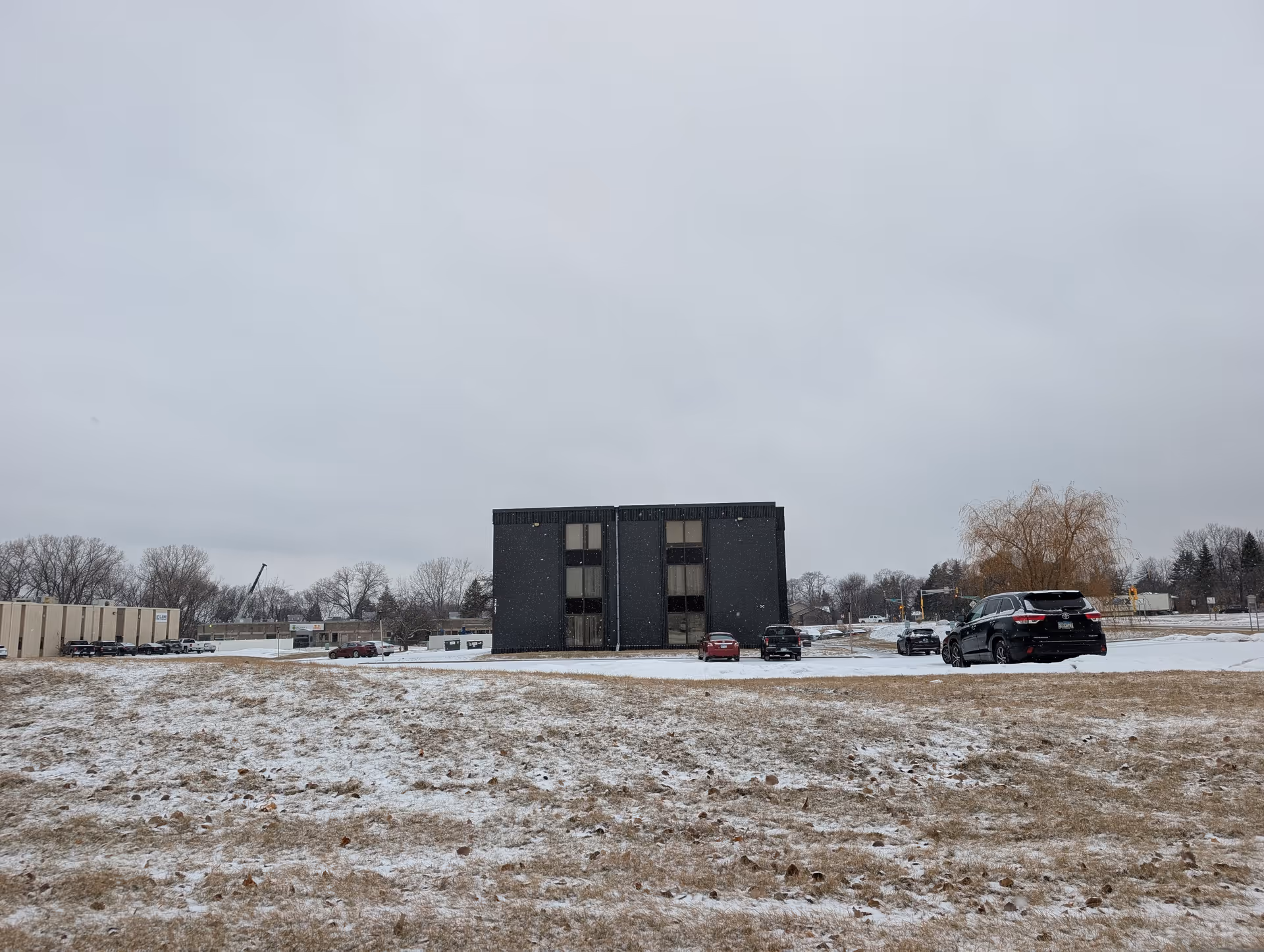Snow-dusted grassy field in front of a three-story dark rectangular building with a few parked cars under an overcast sky.