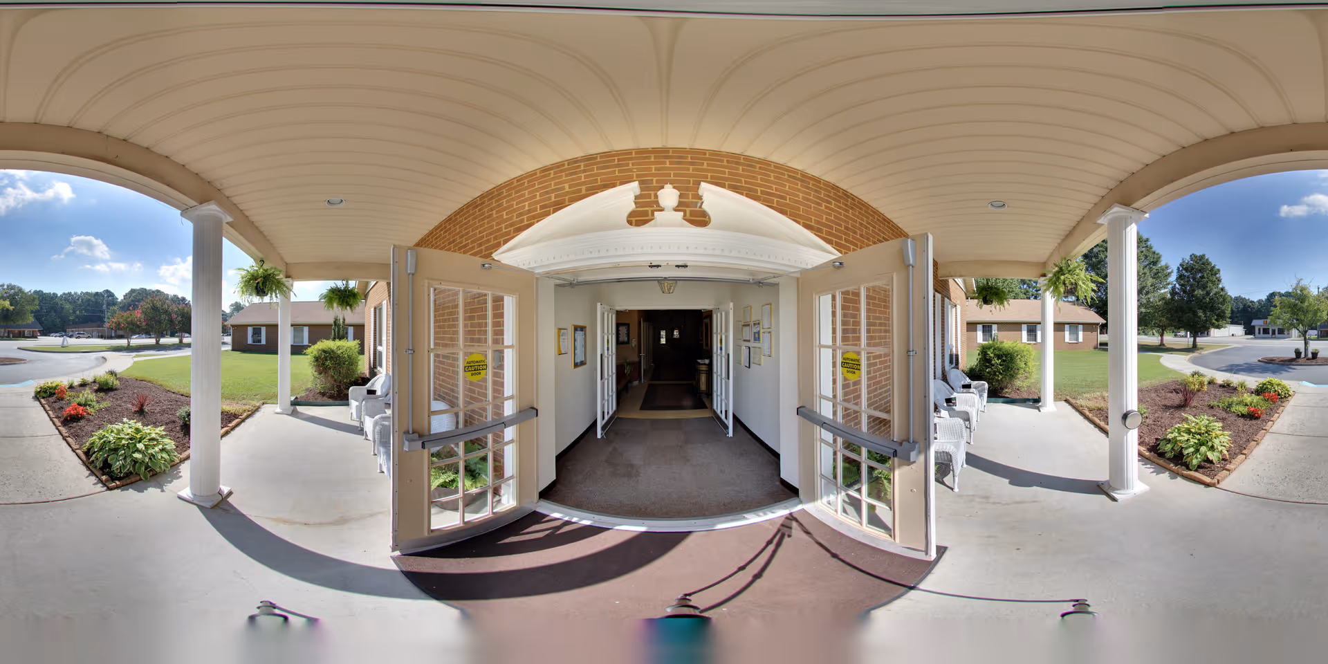 Covered entrance with open double glass doors leading into a brick senior living facility, flanked by white columns, landscaping, and outdoor seating.