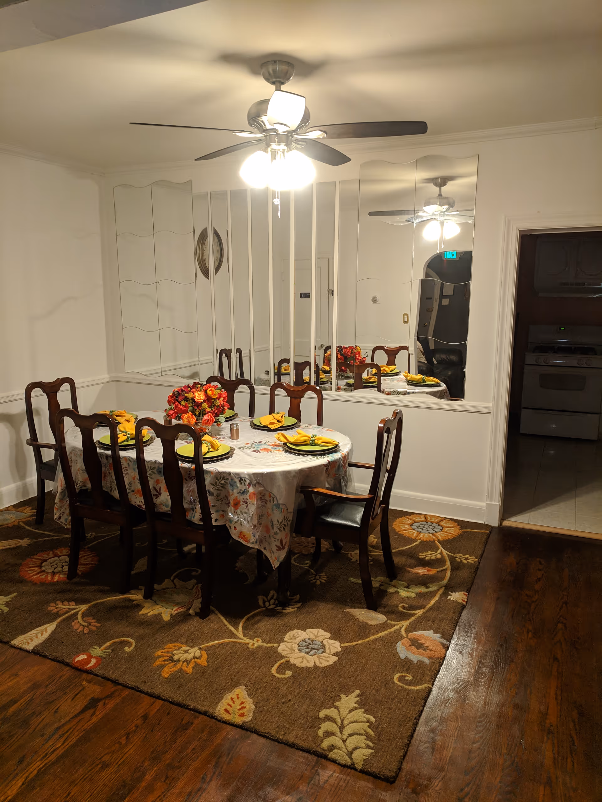 A dining room with a wooden table covered by a floral tablecloth and set with plates, yellow napkins, and a centerpiece of red and orange flowers. Six wooden chairs surround the table, and a ceiling fan with lights is above. The room has a large mirror on one wall and a floral patterned rug on the wooden floor. An open doorway leads to a kitchen area with a stove and refrigerator visible.