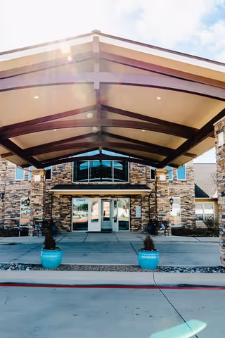 Entrance of a senior living facility with a covered driveway supported by beams, stone facade, glass doors, and two blue planters with small plants on either side of the walkway.