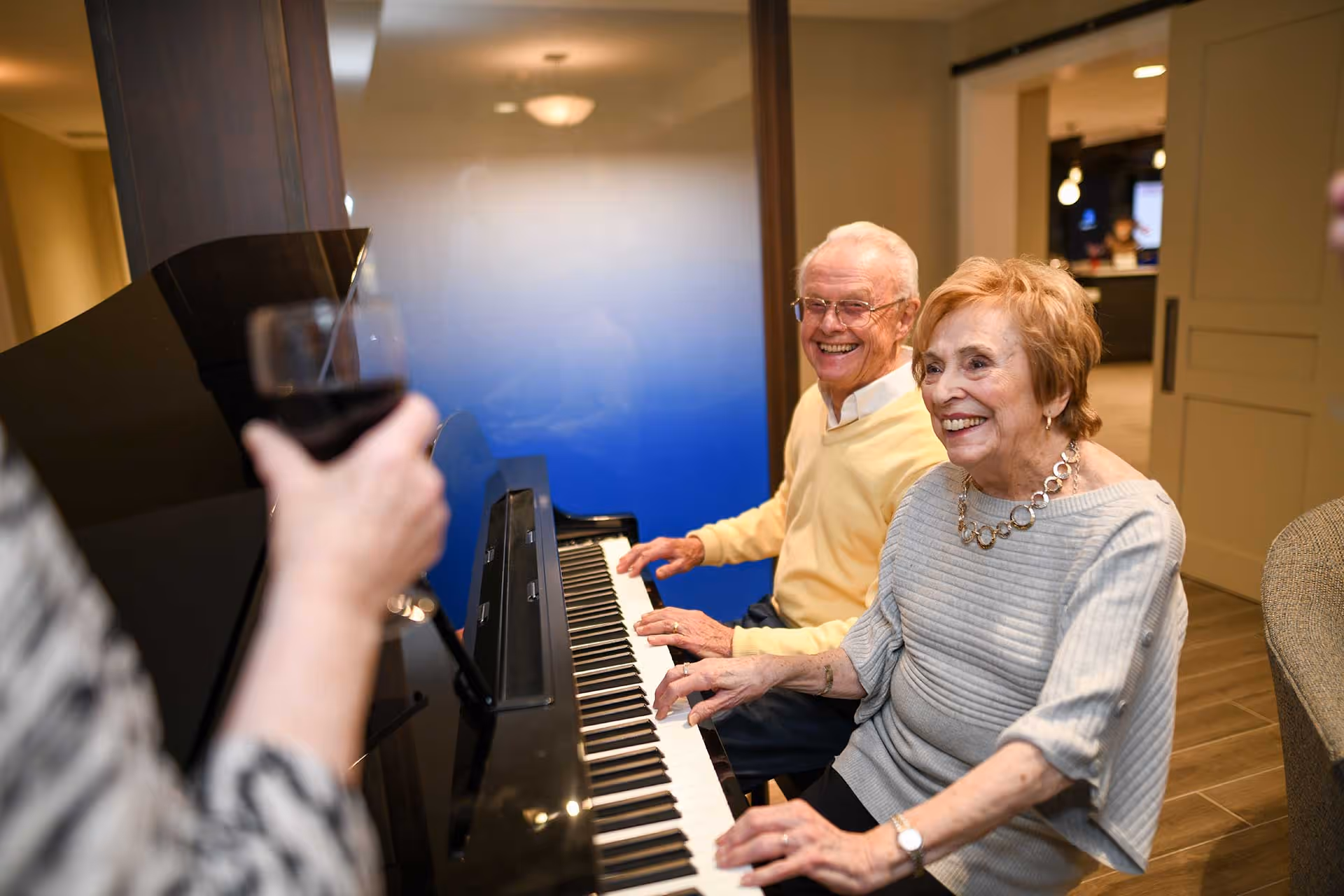 An elderly man and woman sitting at a piano, smiling and playing music together in a well-lit room. A person in the foreground holds a glass of red wine, partially visible. The setting appears warm and inviting with wooden flooring and neutral-colored walls.