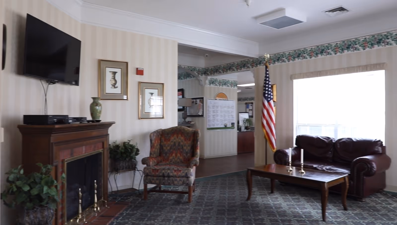 Seated lobby area with an upholstered armchair and leather sofa, coffee table, American flag, wall-mounted TV and fireplace.