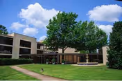 Exterior view of The Residences at Arbor Lake showing a two-story building with large windows, surrounded by green grass, trees, and a clear blue sky with some clouds. There is a paved walkway and a fountain in front of the building.