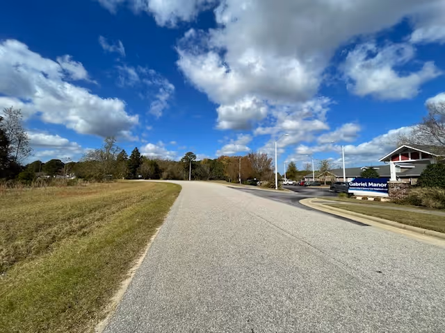 A paved road curves gently to the left with grassy areas on both sides under a partly cloudy blue sky. On the right side of the road, there is a sign for Gabriel Manor Assisted Living Center near a parking area with several cars and a building in the background.