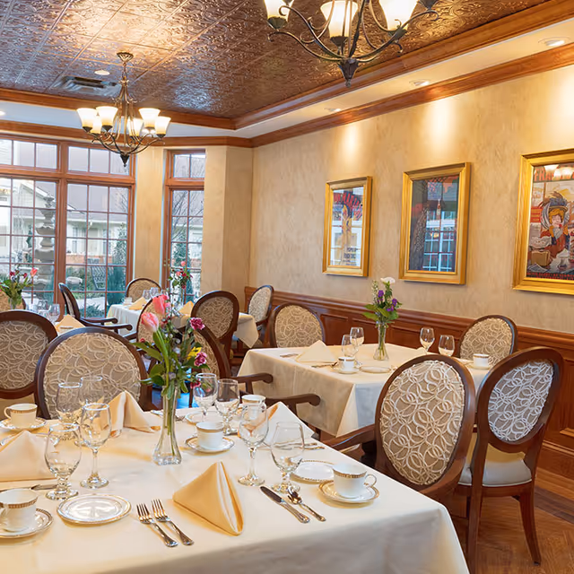 Elegant dining room with multiple set tables, floral centerpieces, upholstered chairs, chandeliers, and framed artwork on the walls.