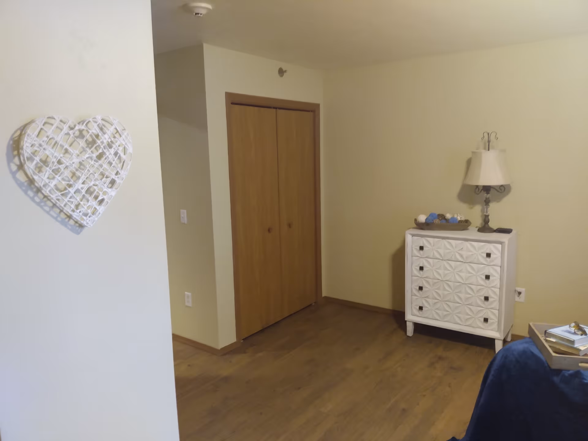 Interior view of a senior living facility room with wooden flooring, a closed wooden closet door, a white decorative chest of drawers with a lamp and decorative items on top, a blue blanket on a piece of furniture, and a white heart-shaped wall decoration.