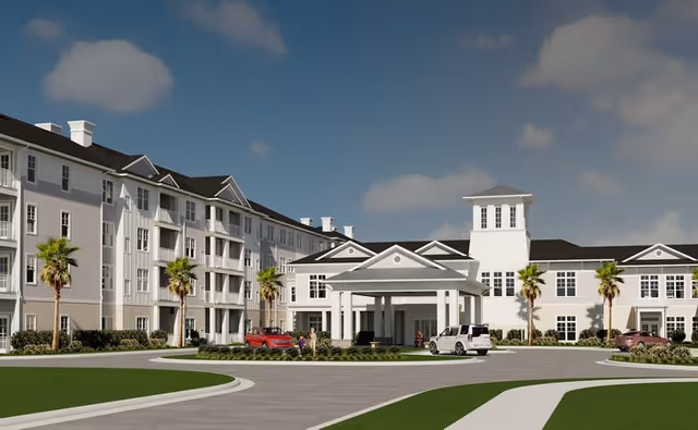 Exterior view of a large, white senior living facility building with multiple floors, palm trees lining the driveway, and several cars parked near the entrance under a covered drop-off area. The sky is clear with a few clouds.