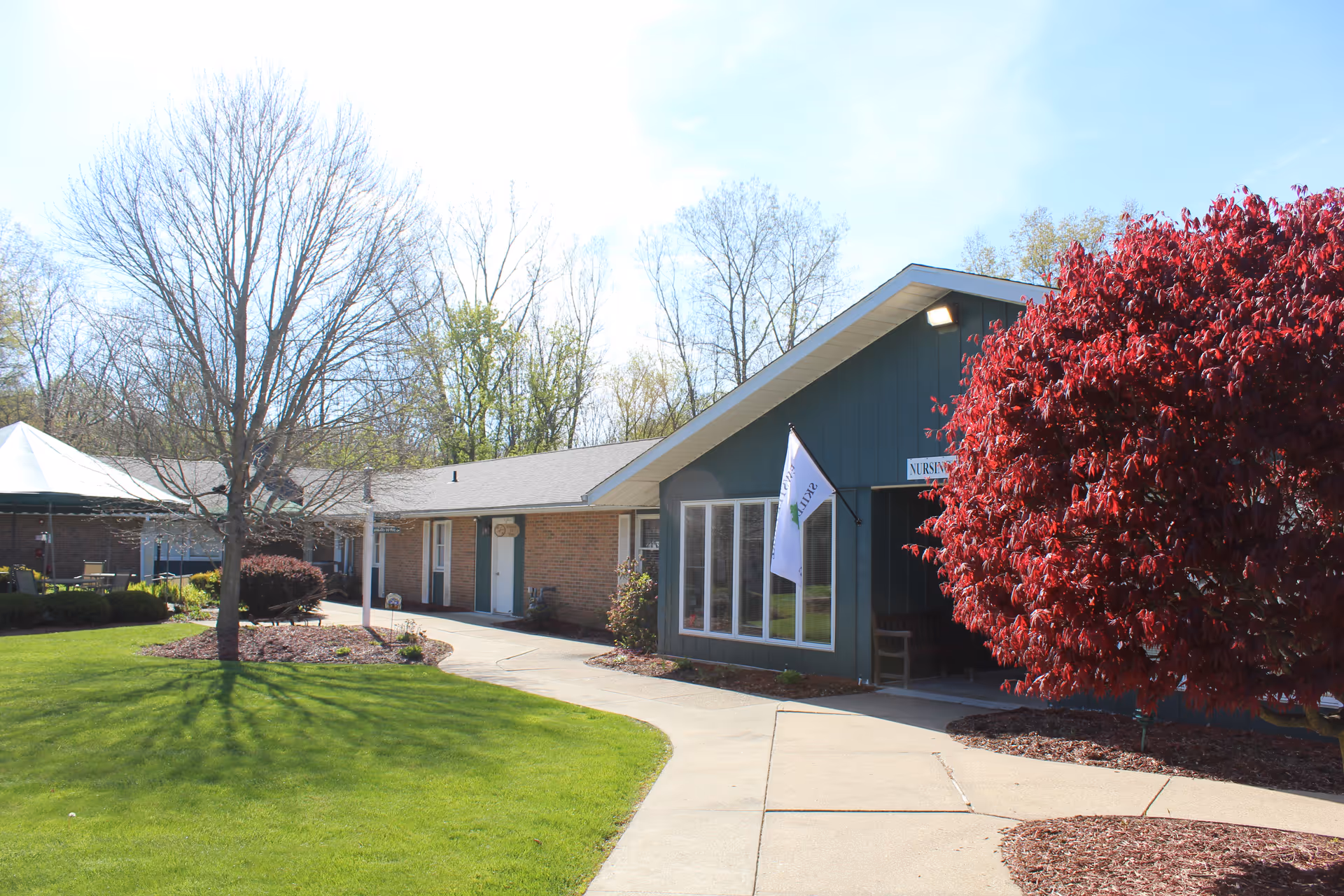 Exterior view of a single-story building with a green lawn and a concrete walkway leading to the entrance. There is a large red-leaved tree on the right side and a white flag near the entrance. Leafless trees and a canopy-covered seating area are visible in the background.