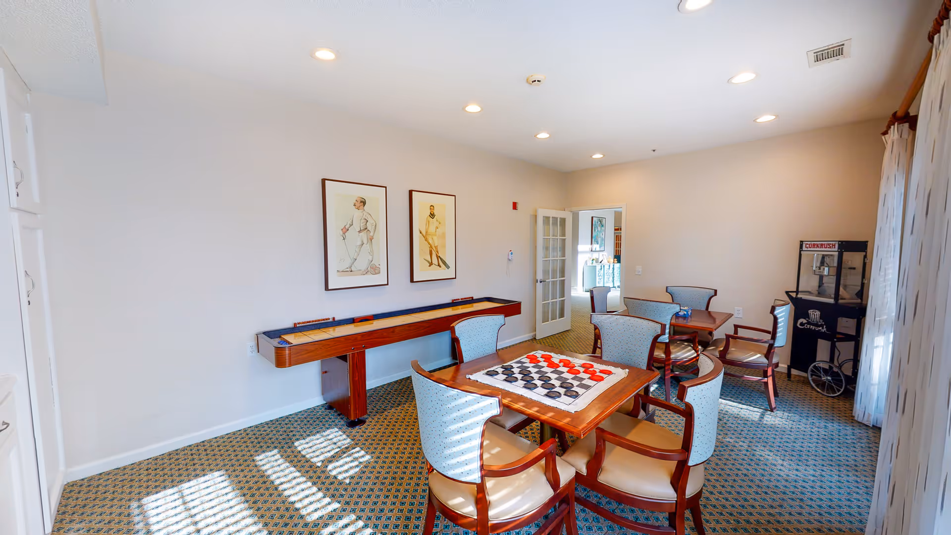 A bright game room with a table set up for checkers, surrounded by four upholstered chairs. In the background, there is a shuffleboard table against the wall with two framed vintage-style sports prints above it. Another table with chairs is visible further back, and a popcorn machine labeled 'Corkrush' stands near a window with sheer curtains. The room has recessed ceiling lights and patterned carpet flooring.