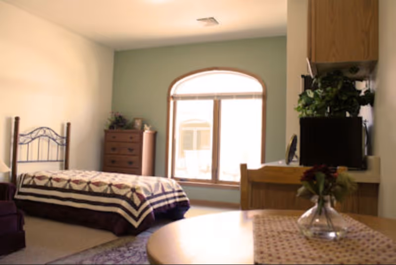 A cozy bedroom with a single bed covered with a patterned quilt, a wooden dresser in the corner, a large arched window letting in natural light, and a small table with a vase of flowers in the foreground. There is also a kitchenette area with a microwave and some plants on the right side.