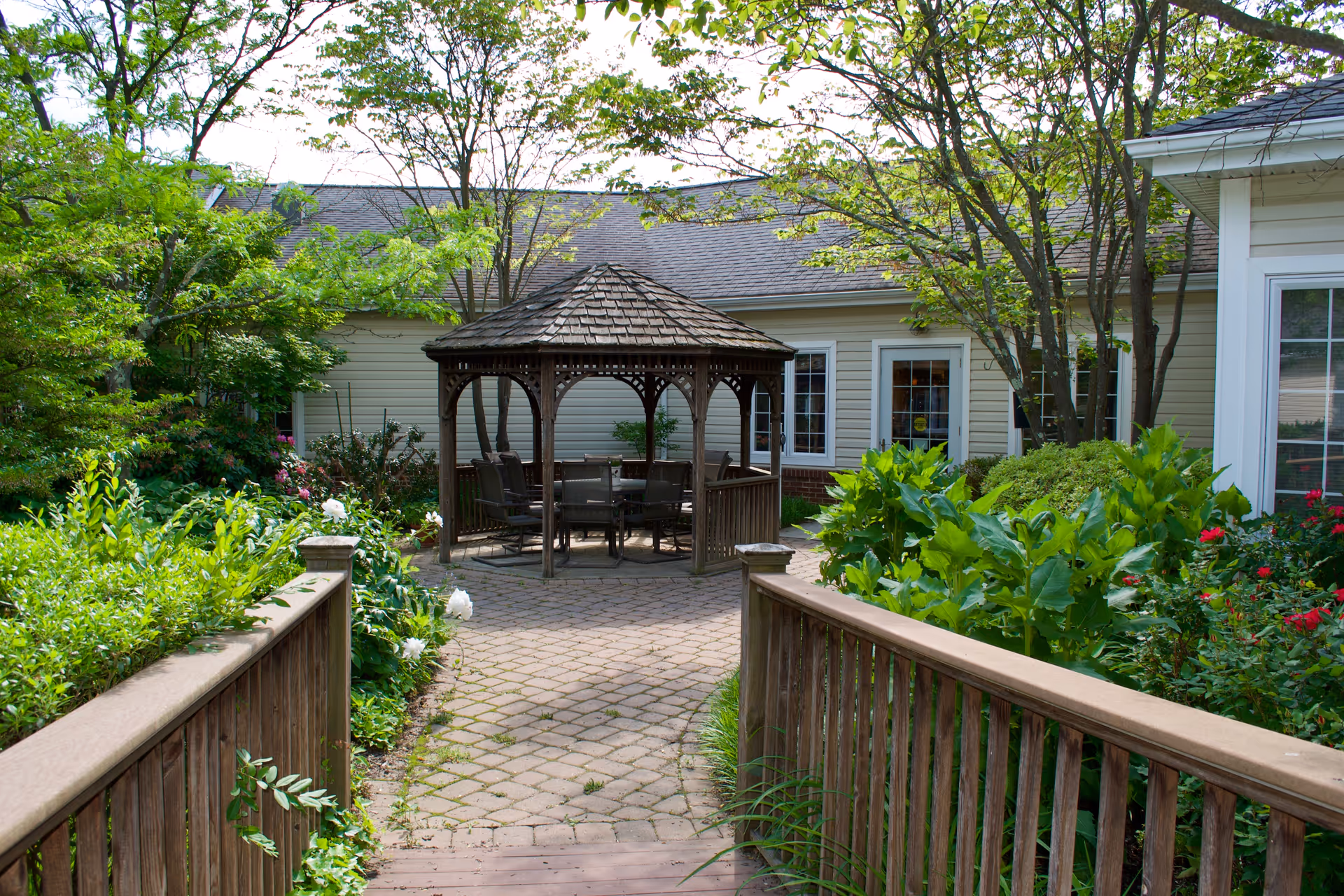 A peaceful outdoor garden area at Barclay Friends featuring a wooden gazebo with chairs and a table inside. The gazebo is surrounded by lush green plants, trees, and colorful flowers. A wooden railing borders a paved walkway leading to the gazebo, with a beige building in the background.