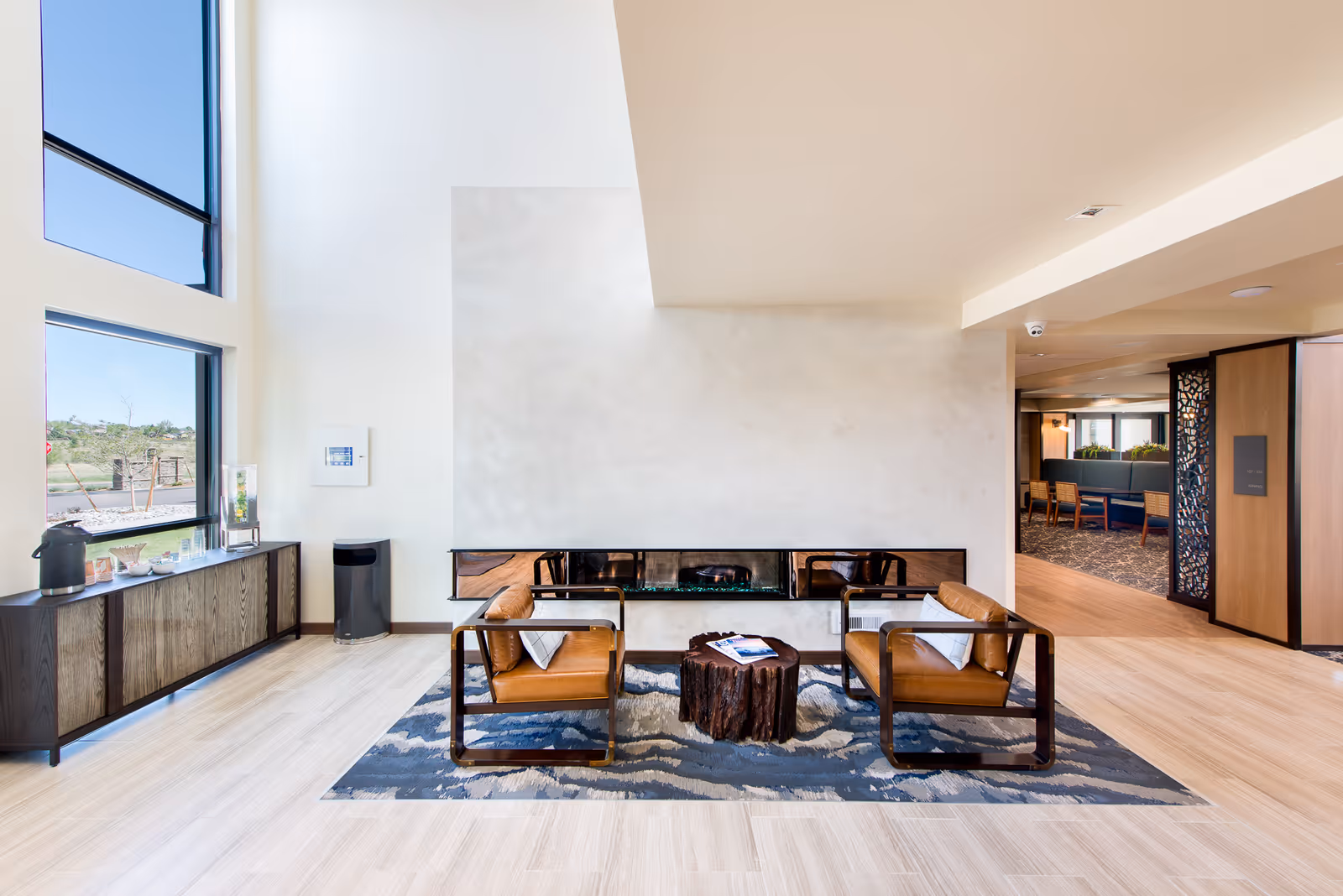 Modern seating area with two brown leather chairs and a small wooden stump table placed on a patterned rug in front of a long, narrow fireplace. Large windows on the left side let in natural light, and a wooden cabinet with a coffee setup is positioned beneath the windows. The space has light wood flooring and a neutral color palette, with an open doorway leading to another room with additional seating.