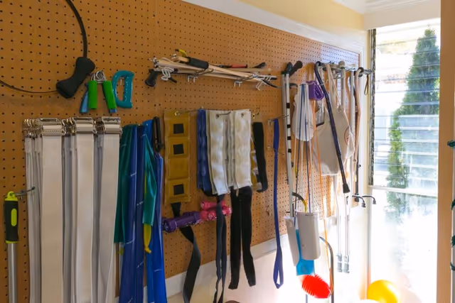 Pegboard wall in a therapy room holding exercise bands, straps, hand weights and walking aids beside a window.