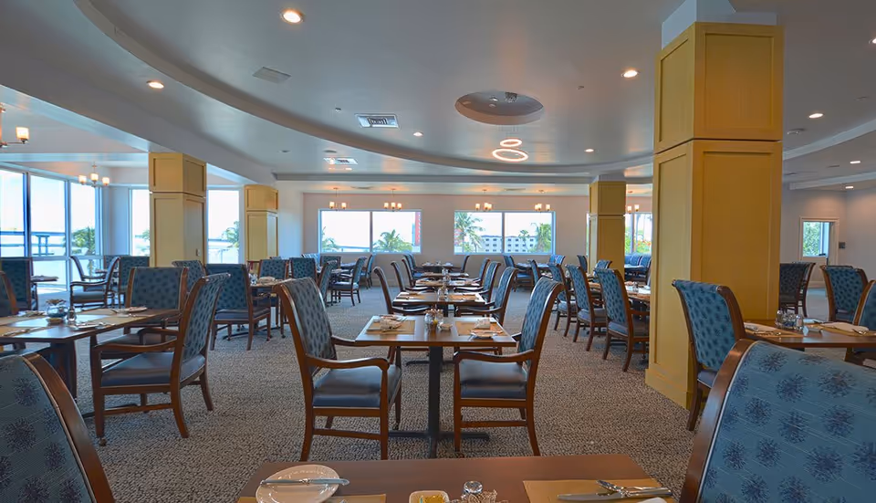 Spacious dining room with multiple tables and chairs arranged neatly. The room has large windows letting in natural light and offering a view of palm trees outside. The ceiling features modern light fixtures and recessed lighting. Tables are set with placemats, plates, utensils, and condiments.