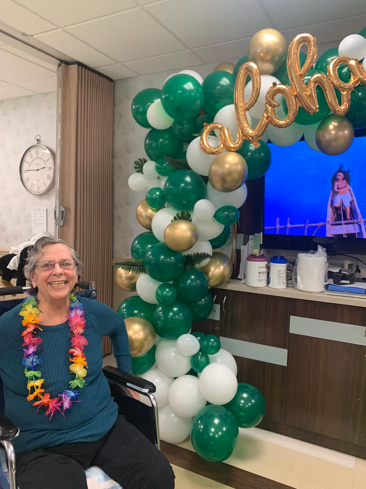 An elderly woman sitting in a wheelchair smiling and wearing a colorful flower lei. Behind her is a decorative balloon arch with green, white, and gold balloons and gold foil letters spelling 'aloha'. A TV screen and cabinets are visible in the background.