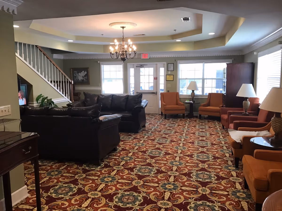 Spacious common room with patterned carpet, leather sofas, multiple armchairs and lamps, a chandelier, and a staircase.