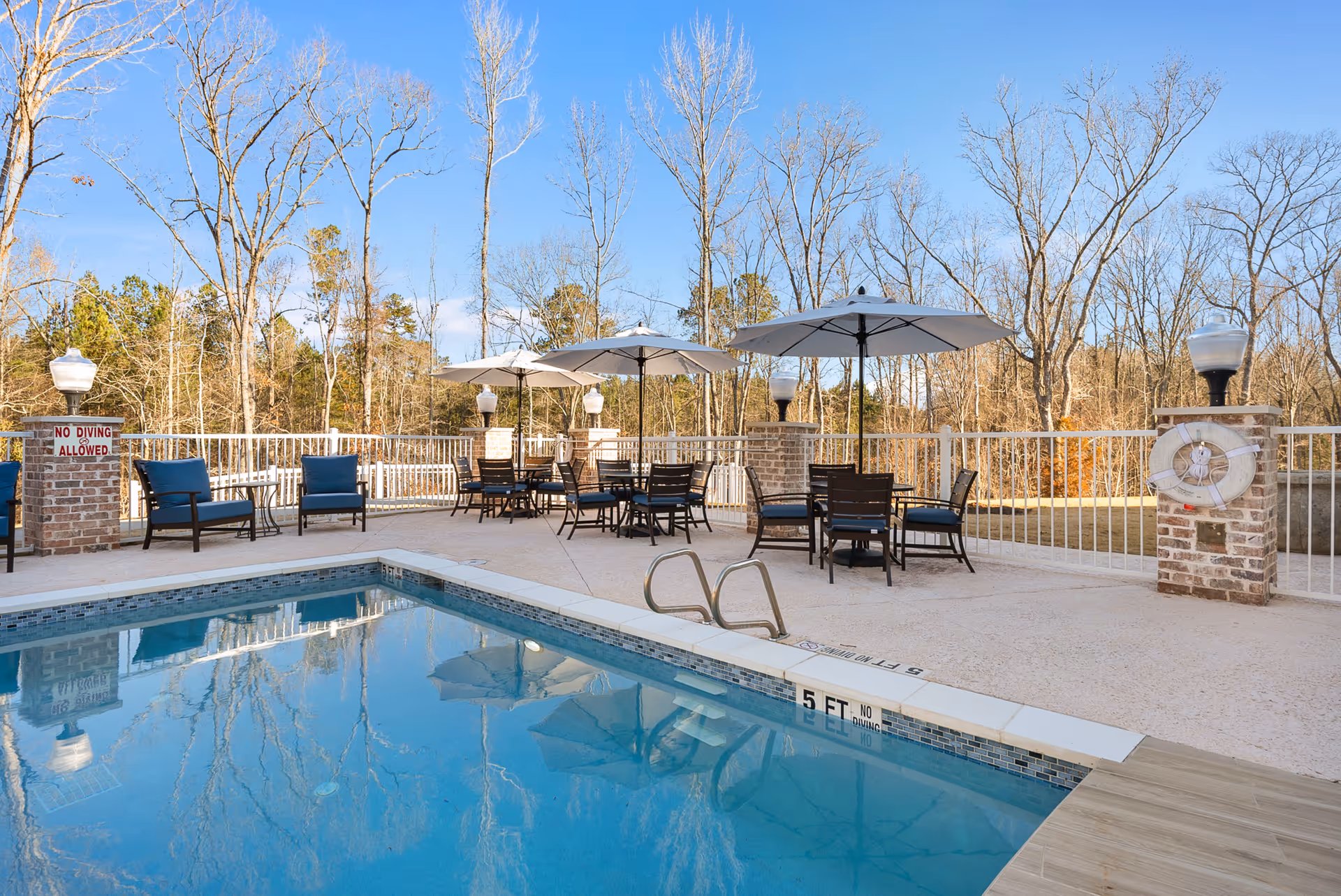 Outdoor swimming pool area with clear blue water, surrounded by a concrete deck. Several tables with umbrellas and chairs are arranged around the pool. There are brick pillars with lamps and a life preserver attached to one pillar. Trees without leaves are visible in the background under a clear blue sky.