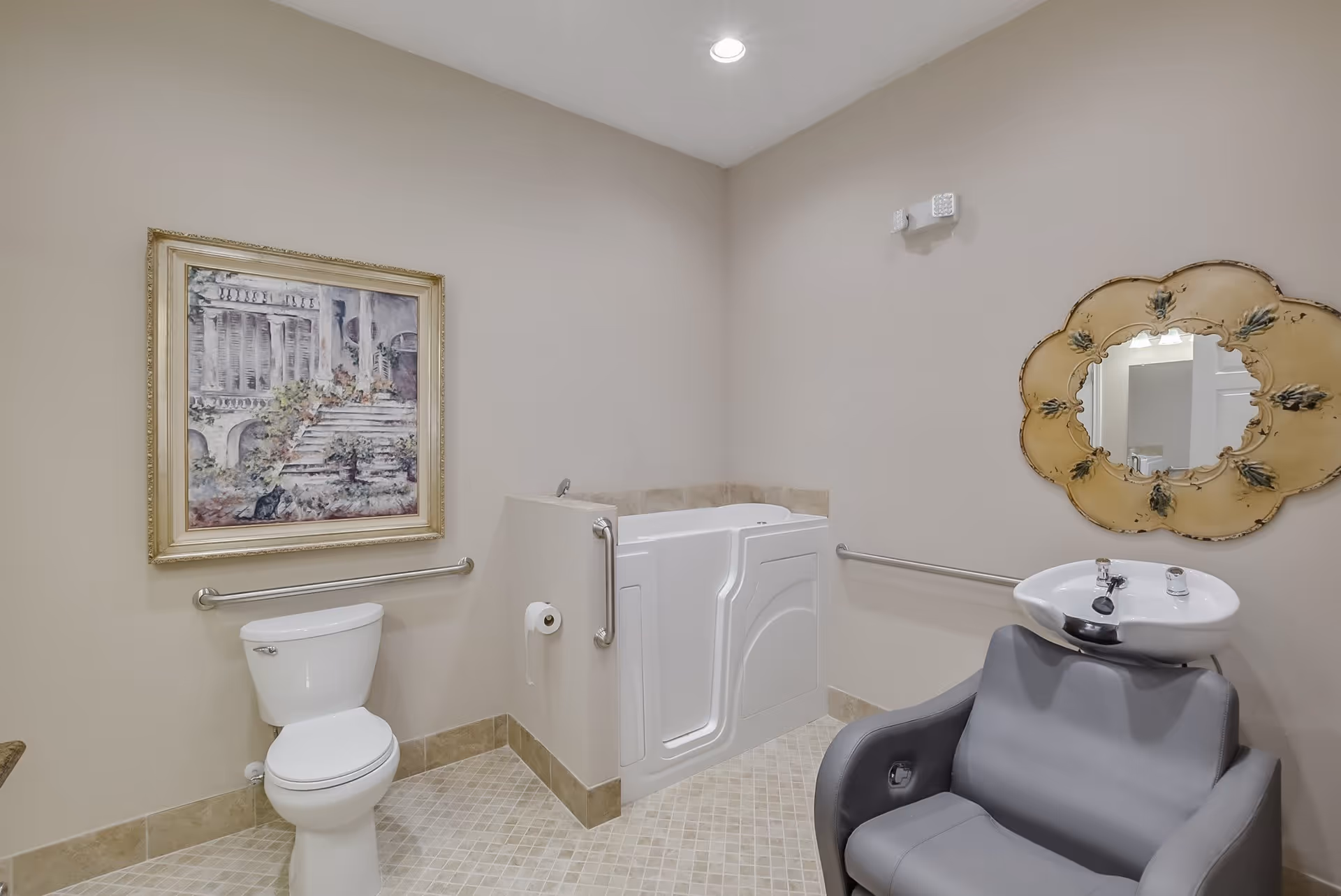 A bathroom with beige walls and tiled floor featuring a white toilet, a walk-in bathtub with grab bars, a gray salon chair with a white sink attached, a decorative wall mirror with a floral design, and a framed painting of a staircase with plants.