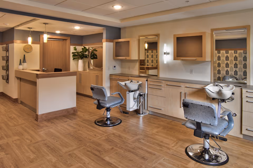 Interior view of a salon area in a senior living facility with two salon chairs in front of sinks and mirrors, wooden cabinetry, a reception desk, and decorative plants and vases.