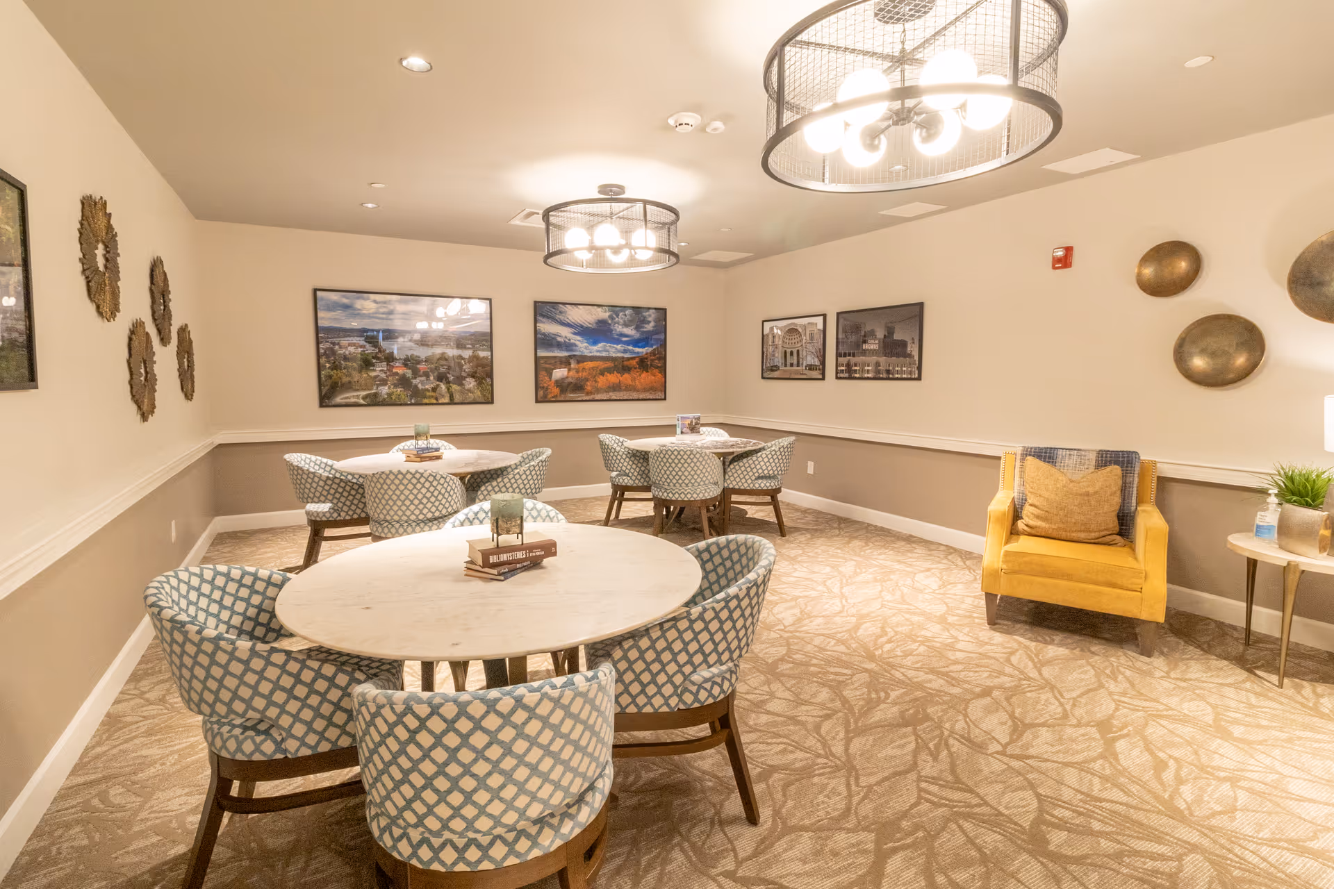 A well-lit interior room with three round tables, each surrounded by four blue and white patterned chairs. The walls are decorated with framed landscape and architectural photographs, as well as metallic wall art. A yellow armchair with a pillow sits in the corner next to a small side table with a plant and hand sanitizer. Two modern circular light fixtures hang from the ceiling.