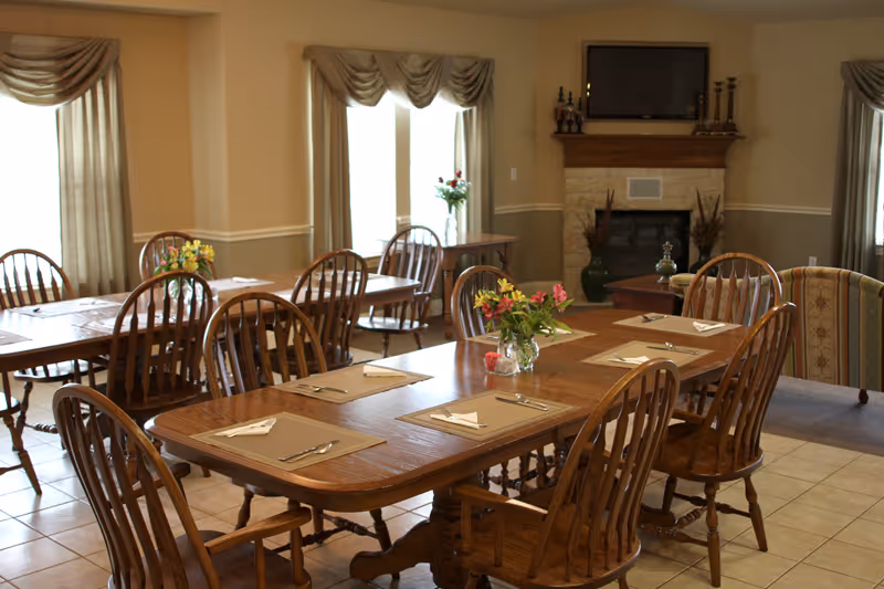 A dining room with wooden tables and chairs set with placemats, napkins, and silverware. There are flower arrangements on the tables, large windows with curtains, and a fireplace with a TV mounted above it in the background.