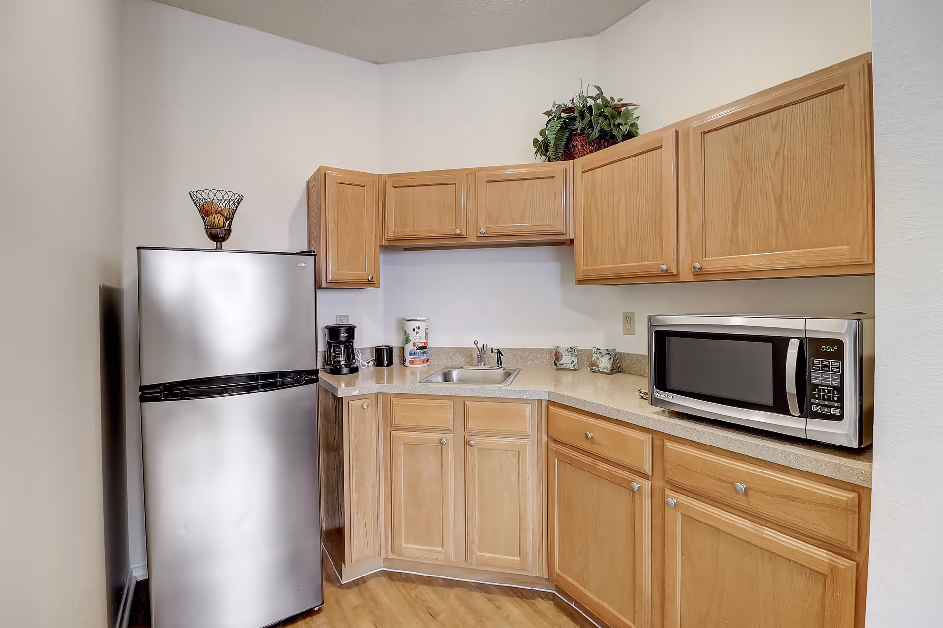 A small kitchen area with light wood cabinets, a stainless steel refrigerator, a coffee maker, a microwave, and a small sink. There is a decorative fruit basket on top of the refrigerator and a plant on top of the cabinets.