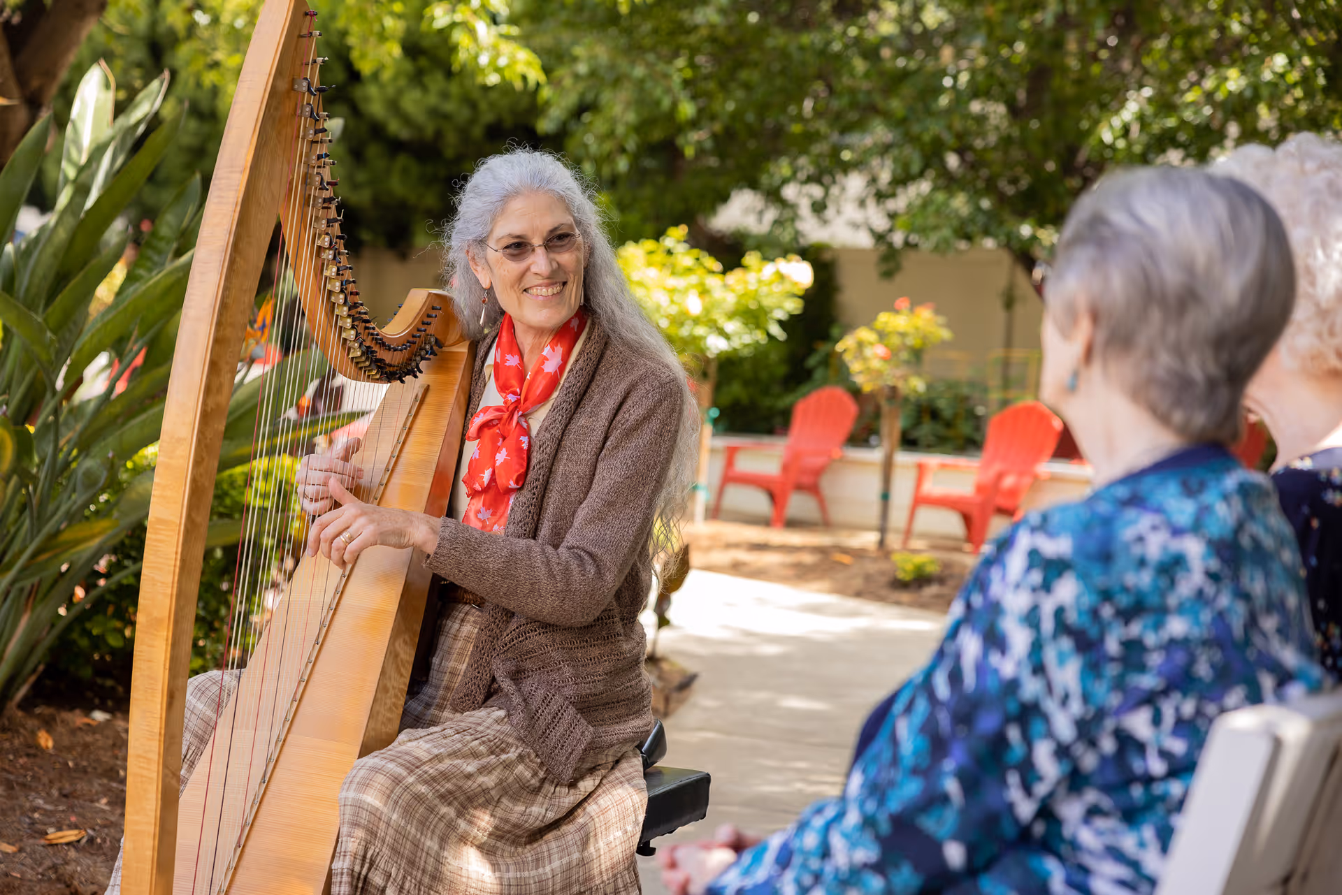 An elderly woman with long gray hair and glasses is playing a wooden harp outdoors in a garden setting. She is smiling and wearing a brown cardigan, plaid skirt, and a red scarf with white maple leaf patterns. Two other elderly women are seated nearby, watching her play. The background features green plants, trees, and red Adirondack chairs.