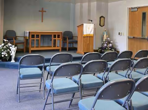 Interior chapel room with a wooden podium, cross on wall, and rows of blue chairs.