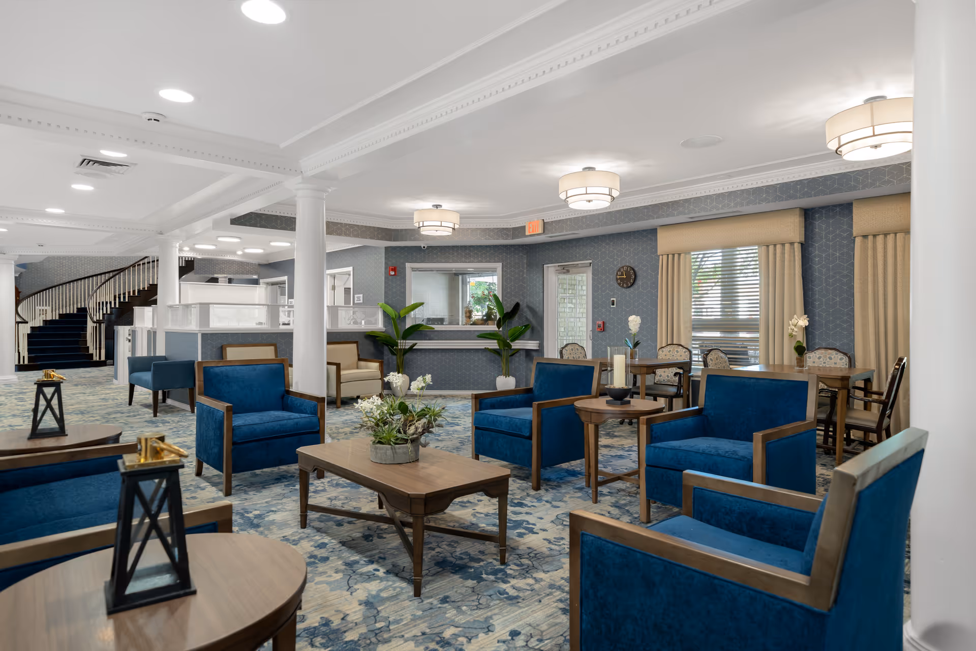 A spacious and elegant common area with blue upholstered armchairs and wooden tables arranged on a patterned carpet. The room features white columns, a staircase with dark carpeting, large windows with beige curtains, and modern ceiling lights. There are also plants and decorative items on the tables, creating a welcoming atmosphere.