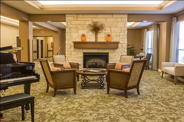 Cozy senior living lounge with a stone fireplace, four chairs, a coffee table, piano, and windows.