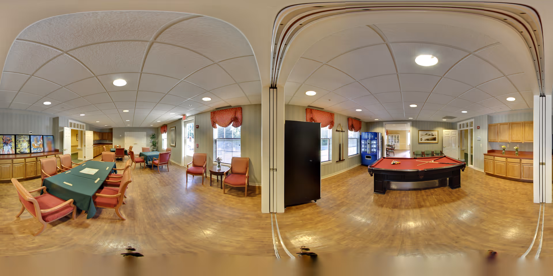 A spacious senior living facility common area with wooden flooring and a white ceiling with recessed lighting. On the left side, there is a long table covered with a green tablecloth surrounded by wooden chairs with red cushions. There are additional seating areas with red cushioned chairs near windows with red valances. On the right side, there is a red felt pool table with cues and balls, a vending machine, and wooden cabinets along the wall. The room is well-lit with natural light coming through the windows.