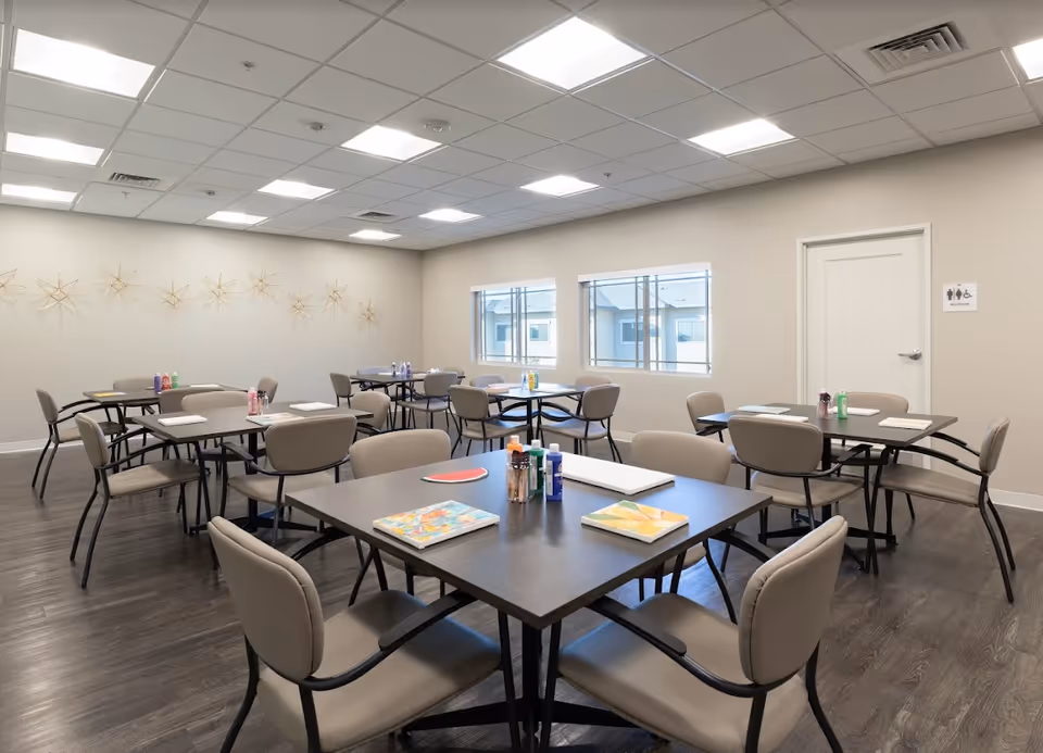 A bright, spacious activity room with multiple tables and chairs arranged for group activities. Each table has art supplies including paint bottles, brushes, and canvases. The room has large windows letting in natural light, a light-colored wall with decorative star-shaped wall art, and a door with a restroom sign.