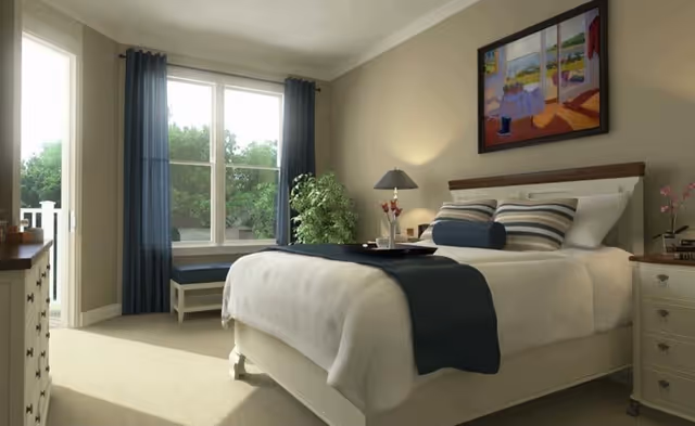 A bright and cozy bedroom with a large bed made with white linens and a dark blue throw blanket. The bed has multiple pillows including striped and cylindrical ones. There is a bedside table with a lamp and a small plant, a framed painting above the bed, a window with dark blue curtains letting in natural light, a cushioned bench under the window, and a dresser on the left side of the room.