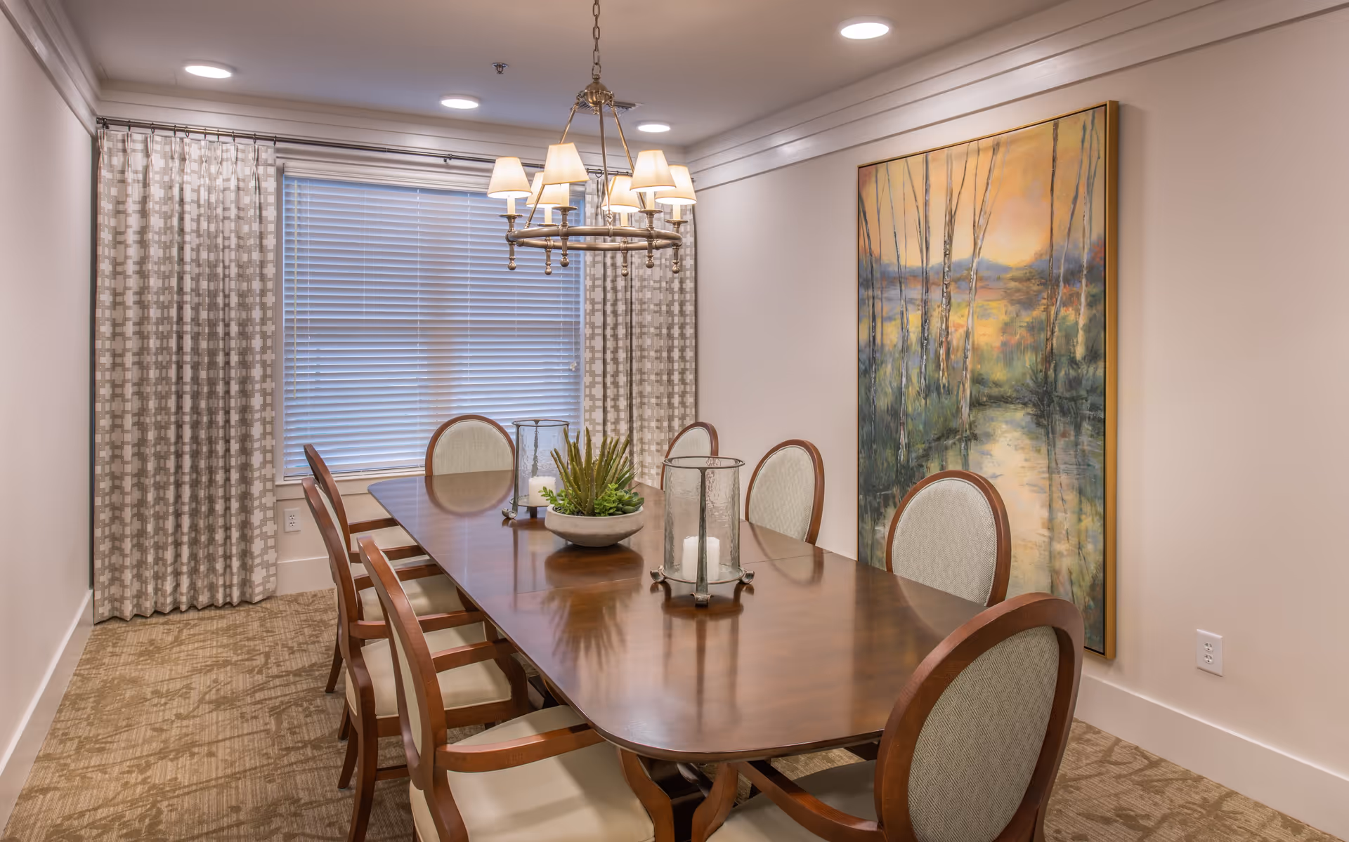 A formal dining room with a long wooden table surrounded by eight upholstered chairs. The table is decorated with a centerpiece of green plants and two glass candle holders. A large window with patterned curtains is at the far end of the room, and a chandelier with multiple lamps hangs above the table. A large framed painting of a nature scene with trees and a river is on the right wall.