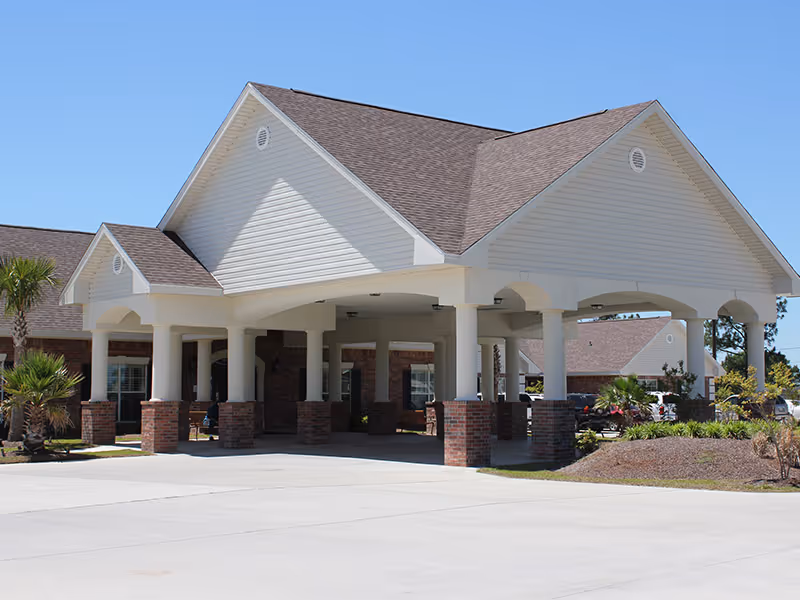 Exterior view of a senior living facility entrance with a large covered drop-off area supported by white columns with brick bases, surrounded by landscaping and a clear blue sky.