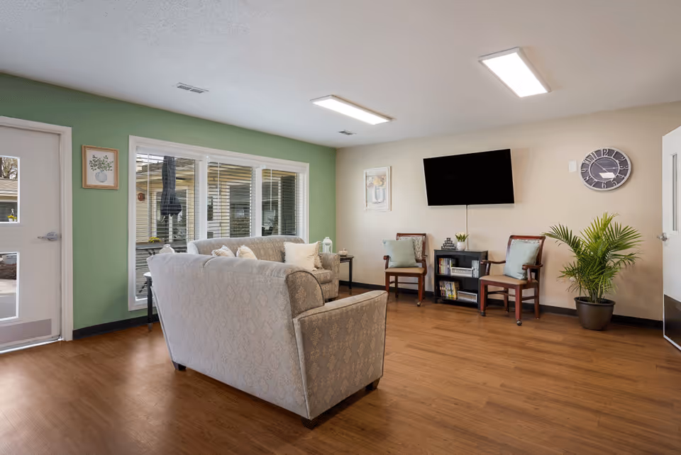 Senior living facility lounge with sofas, chairs, TV, plants, and large windows.