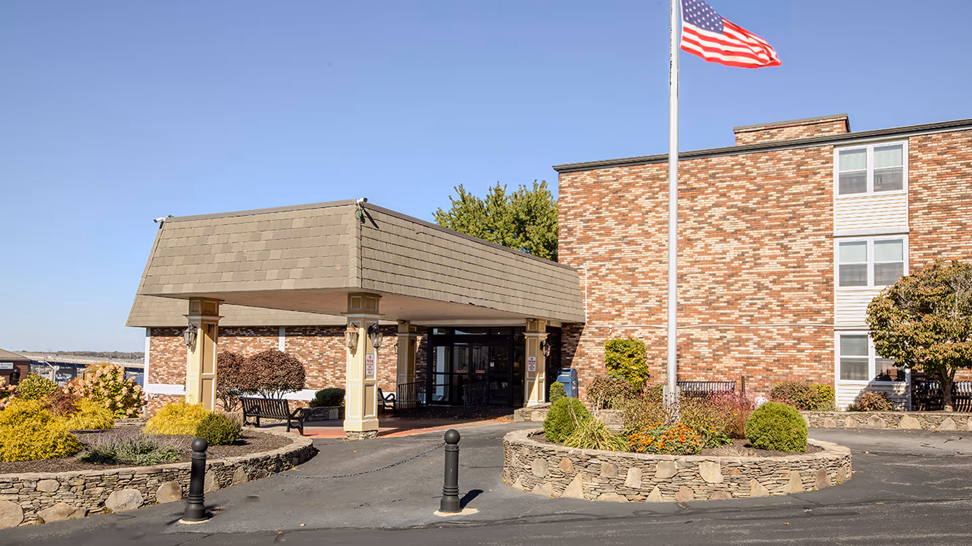 Exterior view of a senior living facility with a brick facade, a covered entrance supported by columns, landscaped garden beds, and an American flag on a flagpole in front of the building under a clear blue sky.