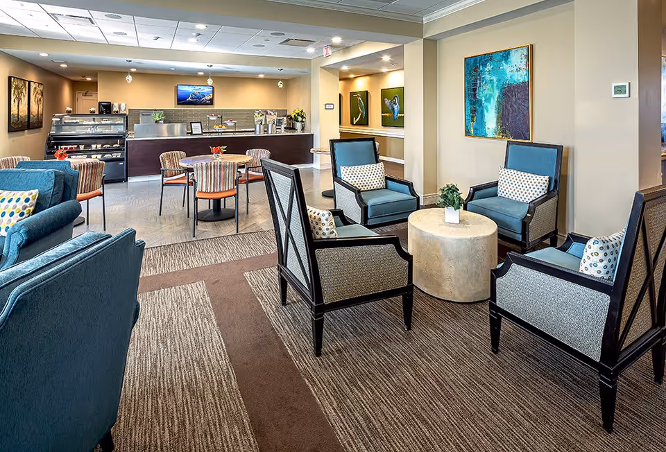 A bright and spacious common area in a senior living facility featuring comfortable seating with blue and patterned armchairs arranged around a round coffee table. In the background, there is a counter with a display case, a television mounted on the wall, and several tables with chairs. The walls are decorated with colorful artwork and the floor is carpeted with a patterned design.