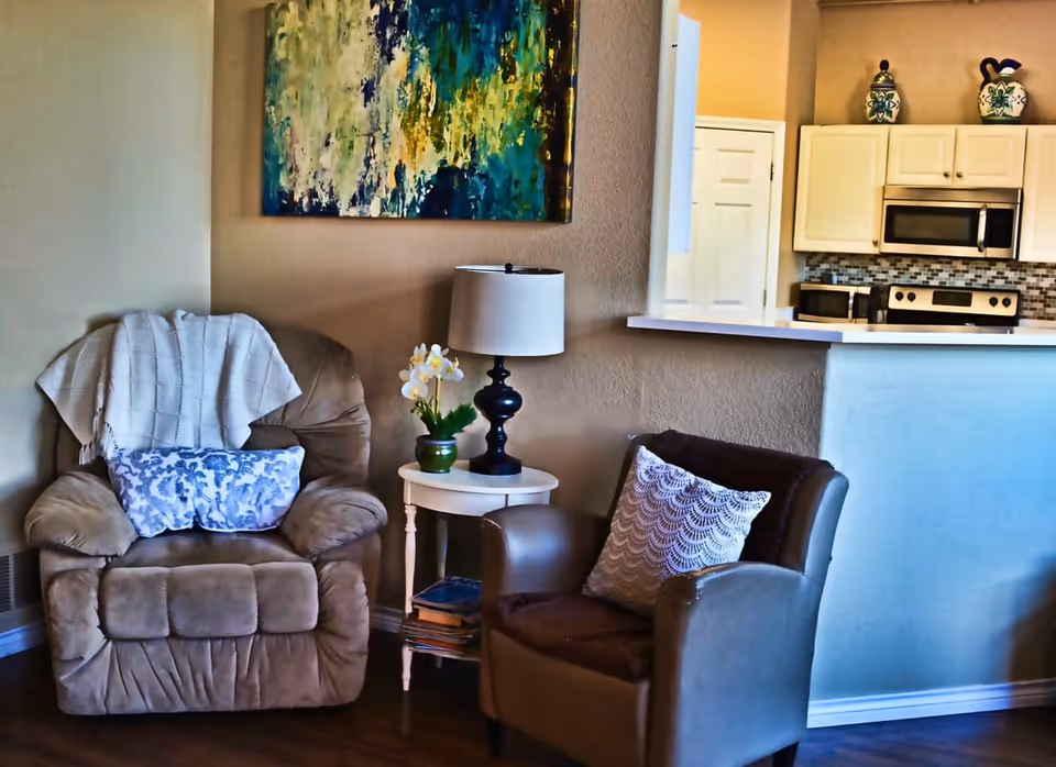 A cozy living area with a beige recliner chair draped with a white blanket and a blue patterned pillow, next to a small white side table holding a black lamp and a small potted plant with white flowers. Adjacent is a brown armchair with a white decorative pillow. In the background, a kitchen with white cabinets, a microwave, stove, and decorative vases on top of the cabinets is visible. A colorful abstract painting hangs on the wall above the recliner.